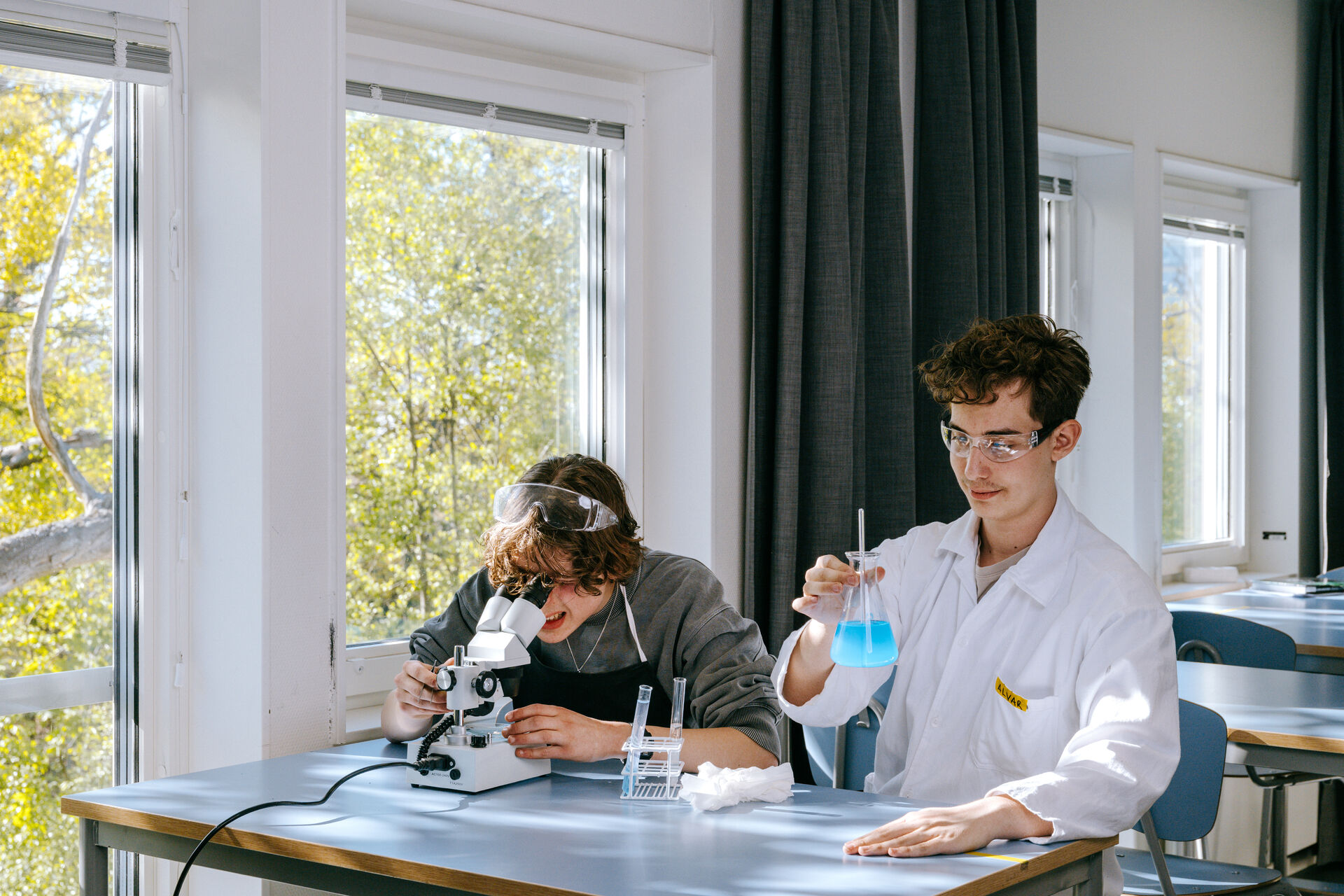 To elever arbeider med naturvitenskapelige eksperimenter i et klasserom i Expandias modulløsning på Ekens skole i Hammarby Sjöstad, hvor den ene eleven bruker mikroskop og den andre blander en blå væske i laboratorieutstyr i et lyst og moderne læringsmiljø.