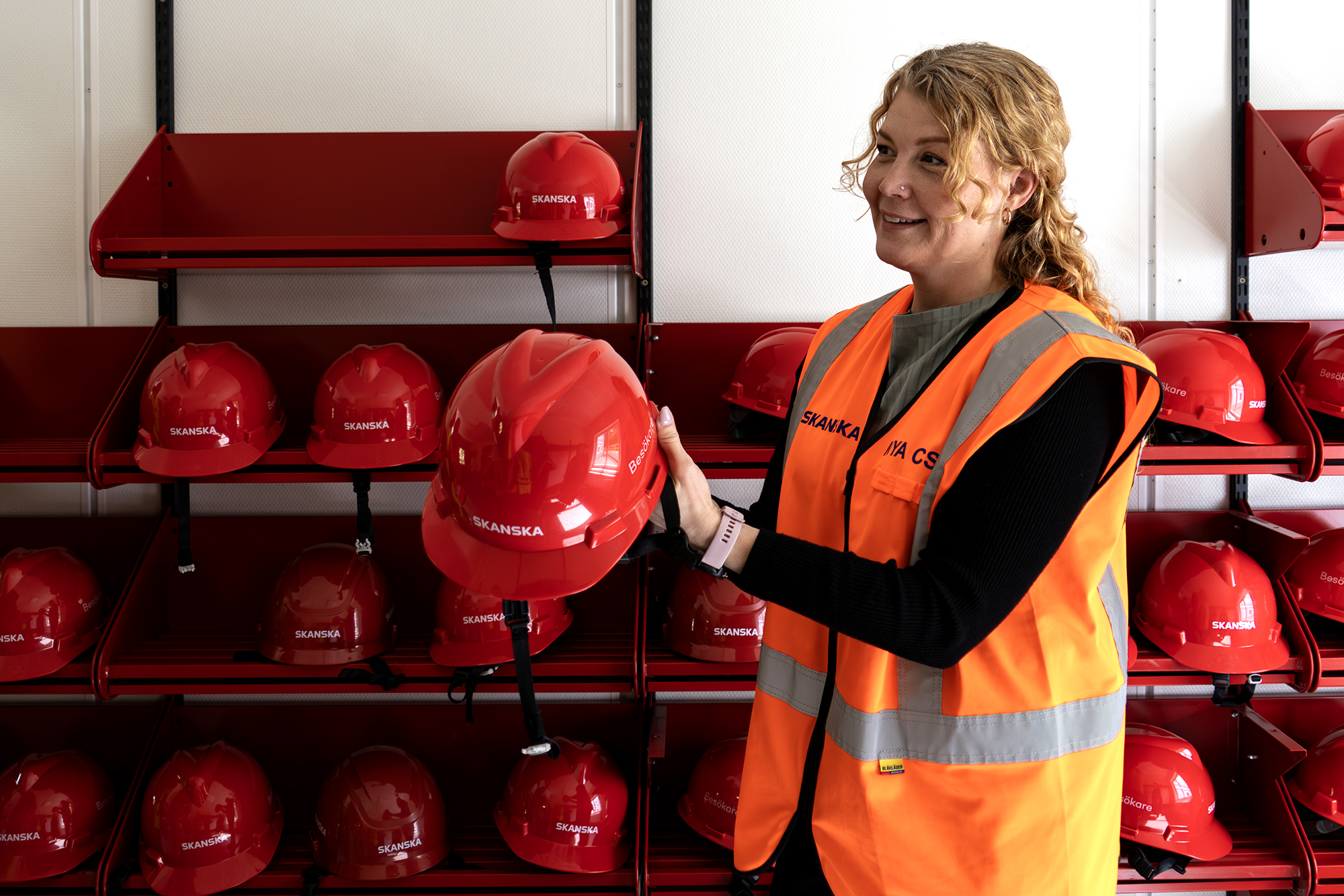 Person i Skanskas projektkontor i Karlstad som tar fram en röd skyddshjälm ur ett ställ. Hjälmarna är prydligt uppställda på hyllor i ett utrymme i kontorsmoduler från Expandia, avsett för besökare och arbetsmiljösäkerhet.