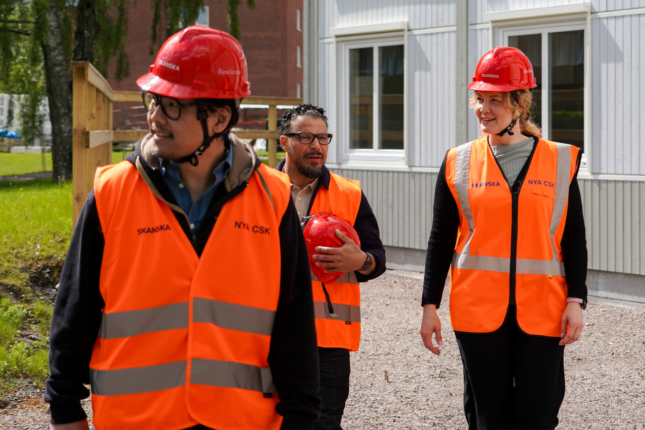 Tre personer i varselvästar och röda skyddshjälmar utanför kontorsmoduler från Expandia vid Skanskas projektkontor i Karlstad. De går tillsammans och samtalar på väg mot arbetsområdet, med modulerna och grön omgivning i bakgrunden.