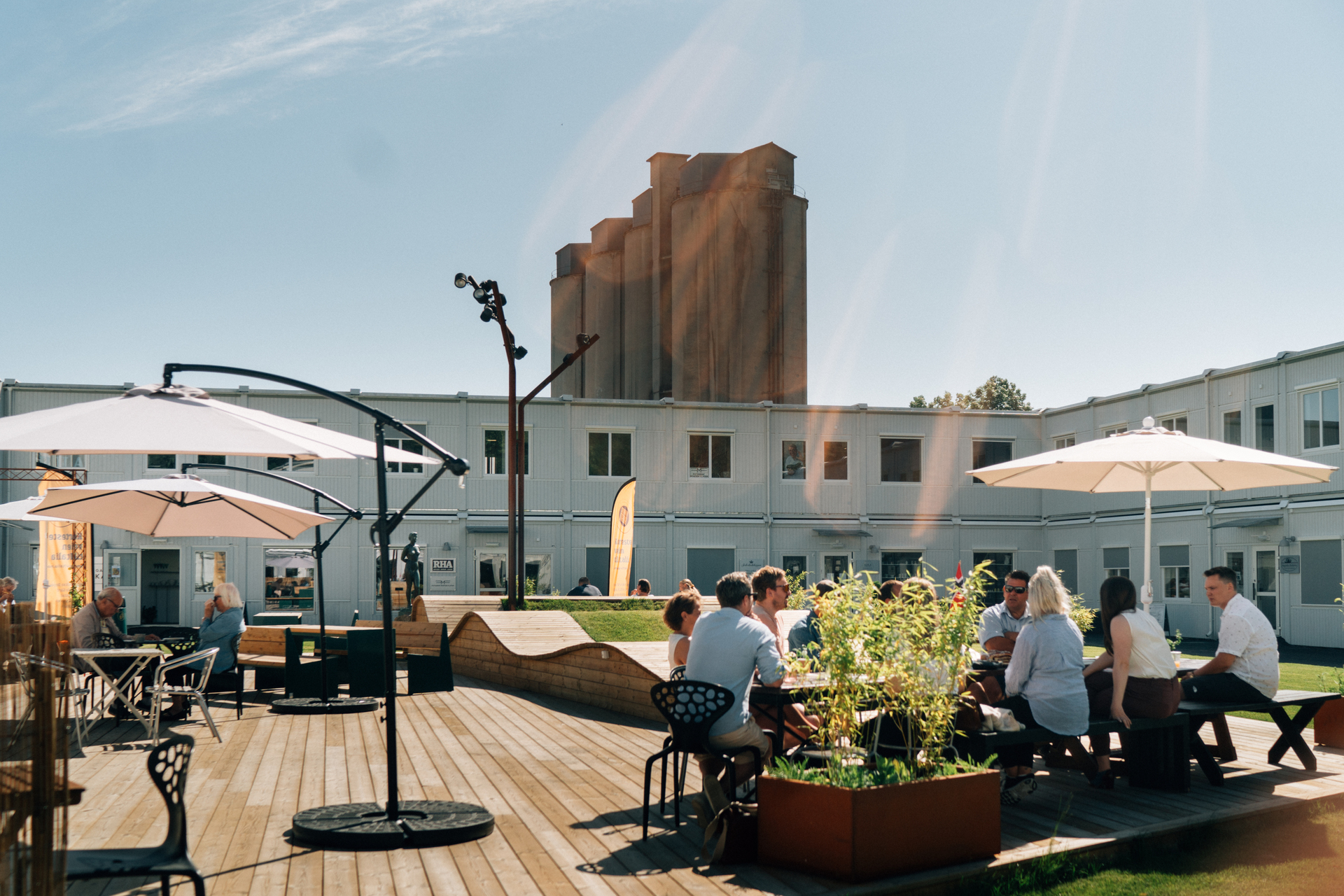 En solrik uteservering med folk som sitter ved bord og sosialiserer på en trebelagt terrasse. Store parasoller gir skygge, og rundt omkring finnes beplantning og sittebenker. I bakgrunnen sees moderne bygg og et høyt industrielt silotårn som reiser seg mot en klar blå himmel. Slemmestad sentrum i Norge.