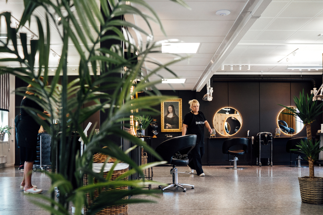 En lys og moderne frisørsalong i Slemmestad sentrum i Norge, med flere stylingstoler og runde speil med belysning langs en mørk vegg. En frisør beveger seg gjennom lokalet, og store grønne potteplanter står i forgrunnen og rundt i rommet. Innredningen er stilren med åpne flater, og i bakgrunnen sees arbeidsstasjoner og salongens utstyr.