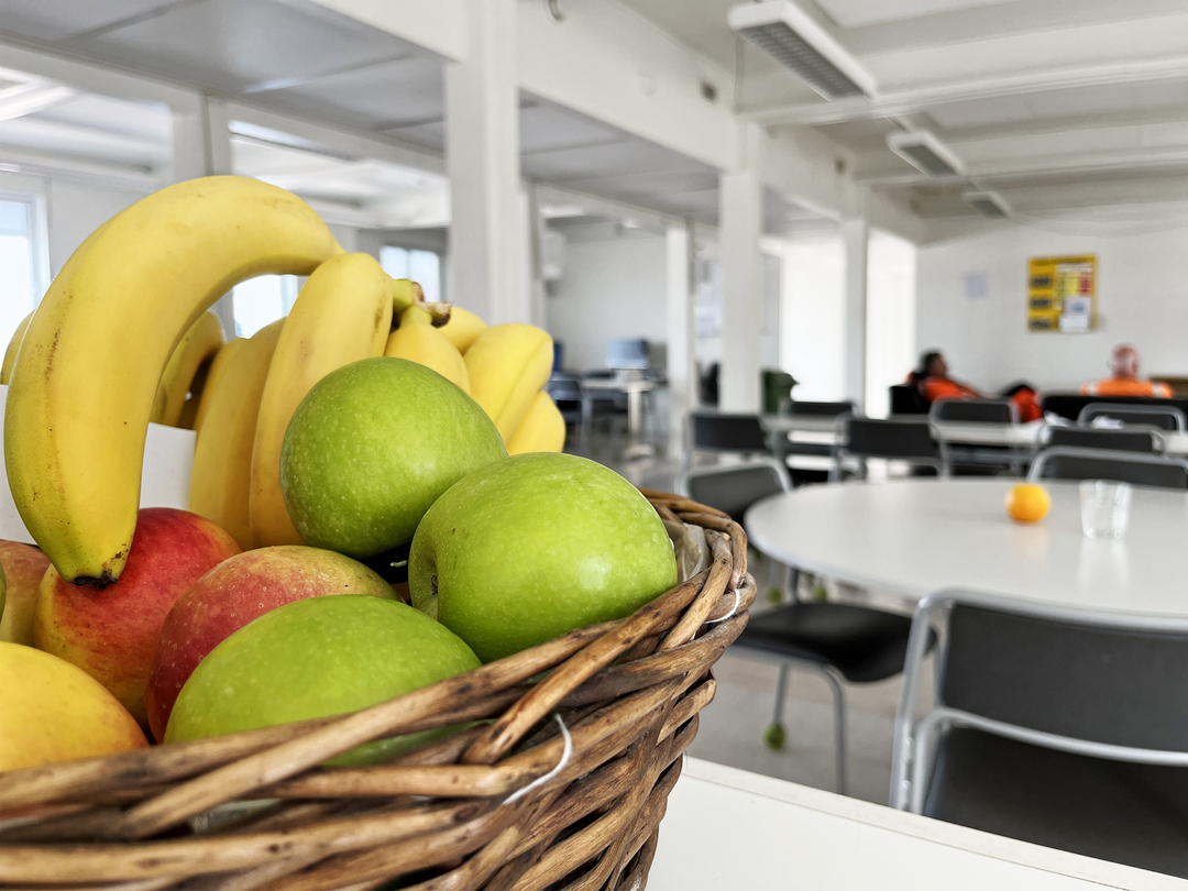 Fruktkorg med bananer och gröna samt röda äpplen i förgrunden, placerad i ett ljust personalrum med bord och stolar; några personer i arbetskläder sitter i bakgrunden.
