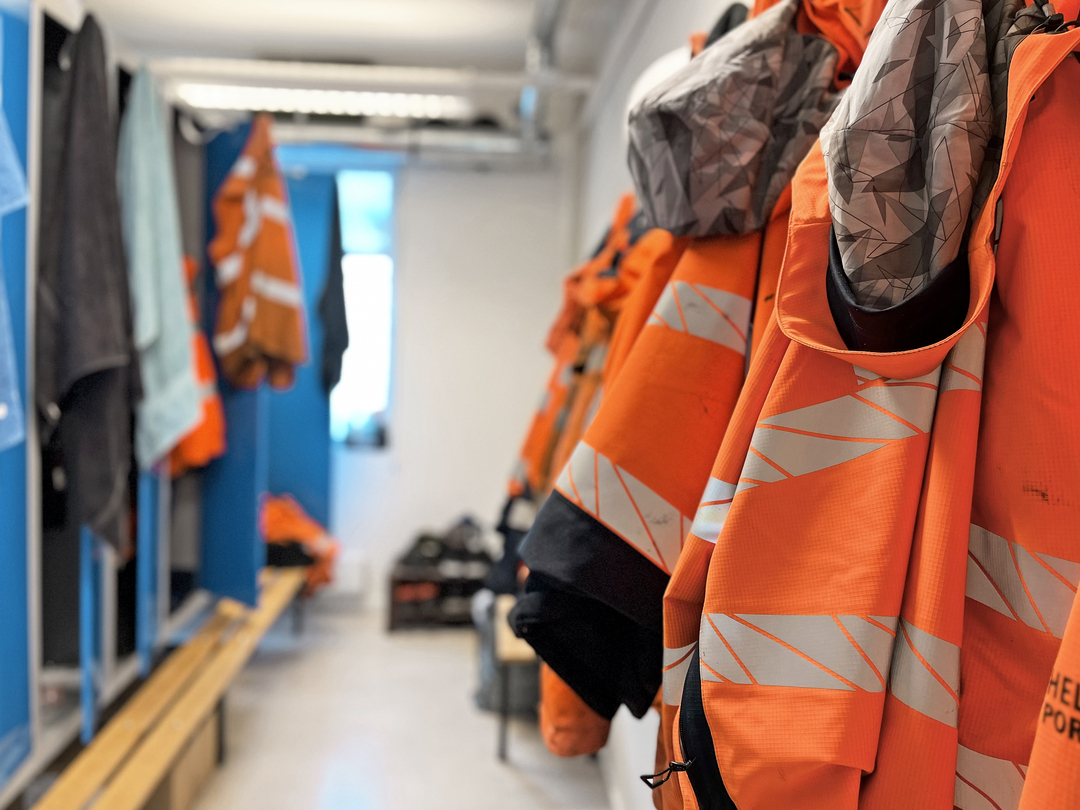 Garderobe med blå skap og benker, hvor oransje varseljakker med refleksbånd henger langs veggen; sko og arbeidsklær sees i bakgrunnen i et lyst og funksjonelt rom.