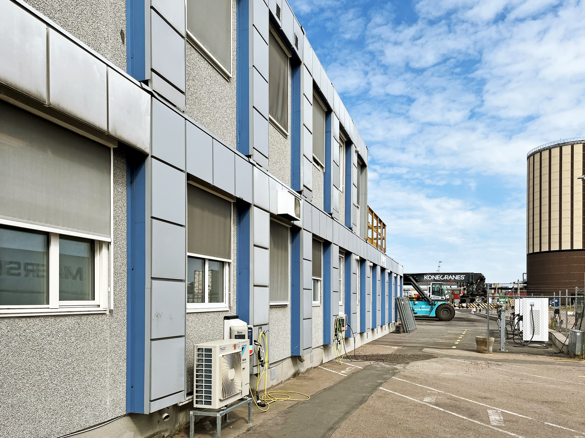 Lang rekke av sammenbygde moduler i Helsingborgs havn i grått, i et industrielt havneområde, med vinduer, eksterne ventilasjonsenheter og et asfaltert område foran; i bakgrunnen sees en høy, sylinderformet industribygning under delvis skyet himmel.