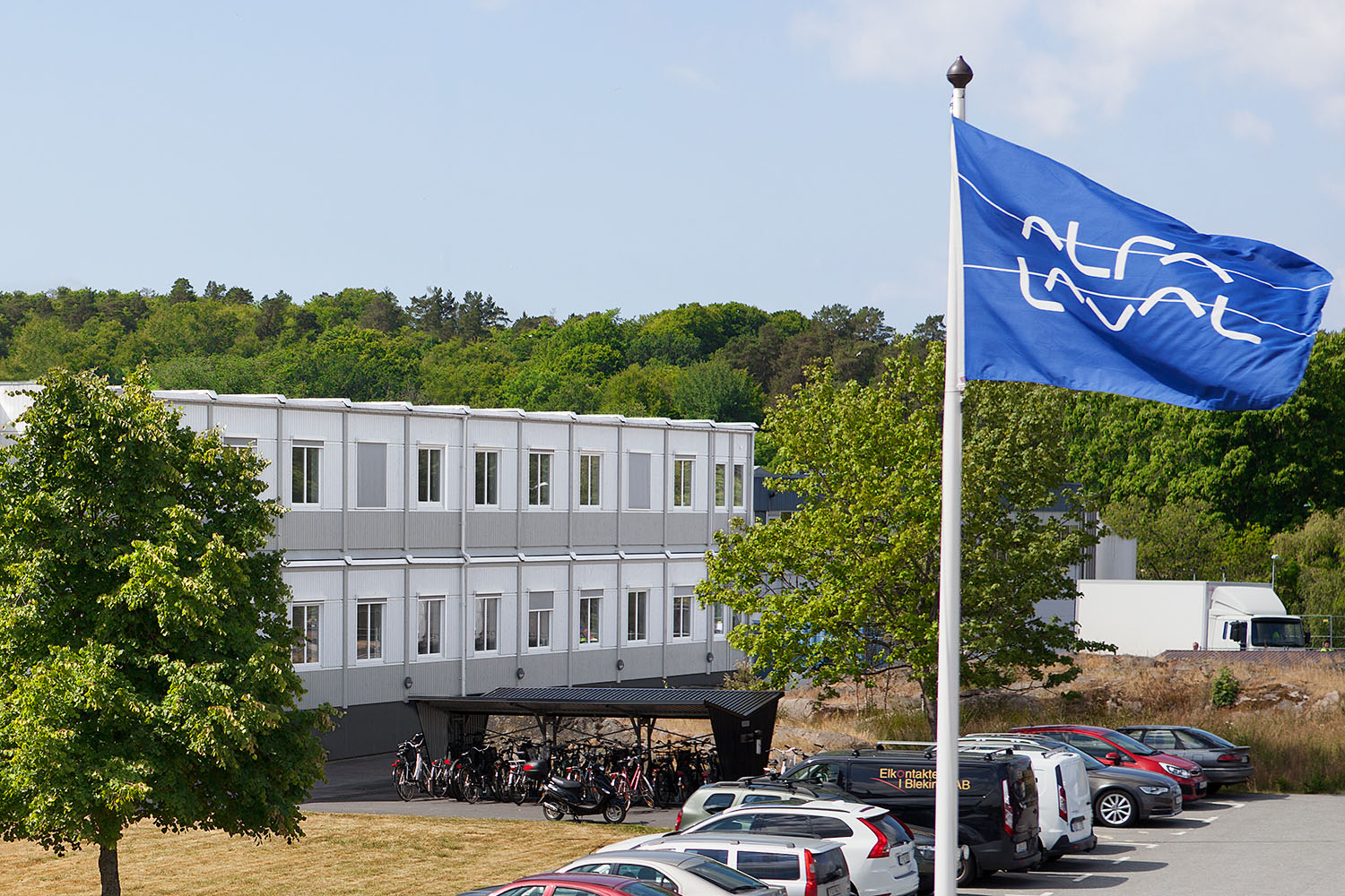 Tvåvånings kontorslösning i ljusgrått placerad i grön omgivning, med parkerade bilar och cyklar i förgrunden samt Alfa Lavals flagga, uppförd med moduler från Expandia.