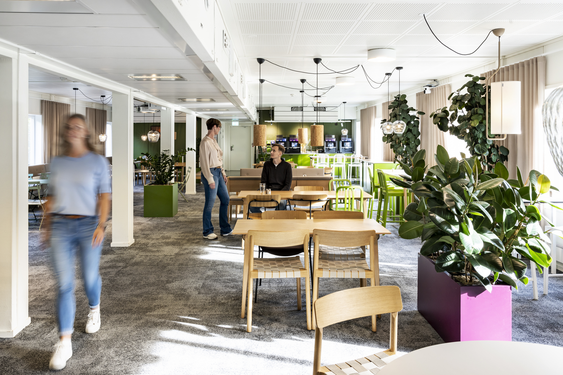 Åpen pause- og møtesone i kontormoduler hos Polypeptide med trebord, grønne stoler, rikelig med planter og pendellamper, hvor ansatte beveger seg og samtaler i et lyst og moderne arbeidsmiljø. Kontormoduler fra Expandia.