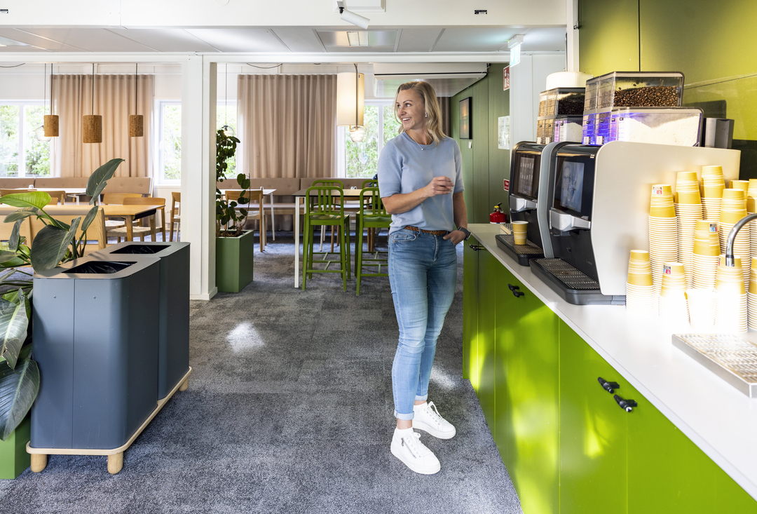 Pauseområde og kaffestasjon i kontormoduler hos Polypeptide med grønne skap, kaffemaskiner, sittegrupper, planter og myk belysning, der en medarbeider tar kaffe i et trivelig arbeidsmiljø – kontormoduler fra Expandia