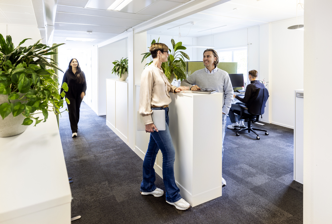 Lyst kontormiljø i kontormoduler hos Polypeptide, hvor ansatte samtaler ved en ståplass, med åpne arbeidsplasser, grønne planter og teppegulv som skaper et moderne og trivelig arbeidsmiljø – kontormoduler fra Expandia.