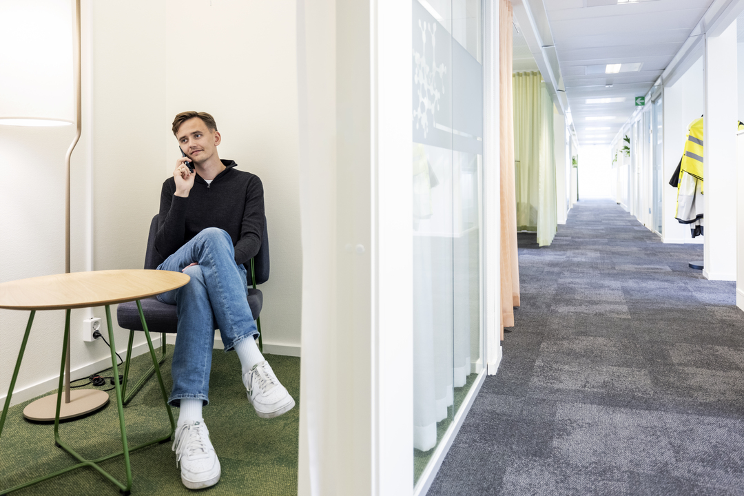 Avskilt sitteområde i kontormoduler hos Polypeptide, hvor en ansatt sitter og snakker i telefon ved et lite bord, ved siden av en glasskorridor med tekstilgulv og myk belysning i et rolig og moderne arbeidsmiljø – kontormoduler fra Expandia.