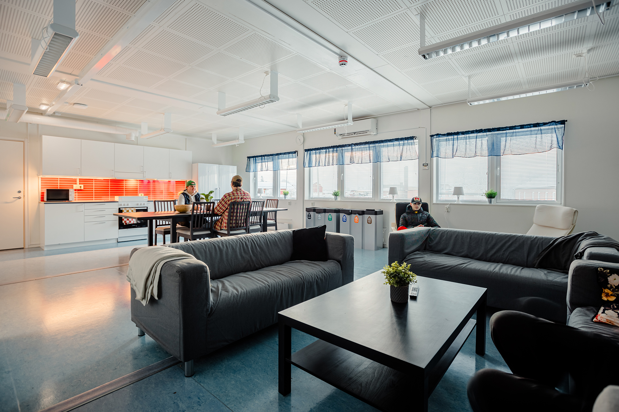 Gemensamt allrum och kök i elevboendemoduler, med soffgrupp i förgrunden och matplats där flera personer sitter tillsammans i en ljus och rymlig miljö.