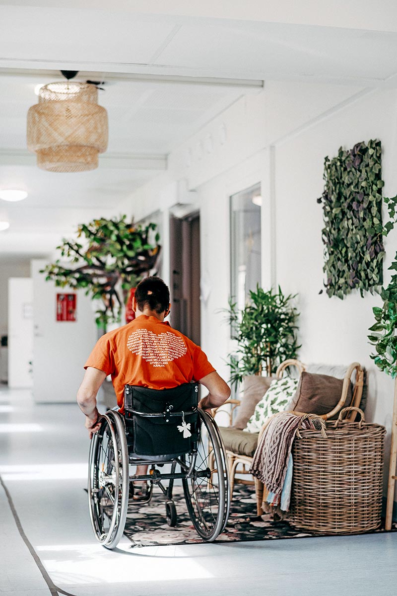 En person i rullstol rör sig genom en ljus och hemtrevlig korridor i Insperia daglig LSS-verksamhet i Ekerö. Miljön är inredd med växter, sittmöbler och mjuka textilier som skapar en varm och välkomnande atmosfär.