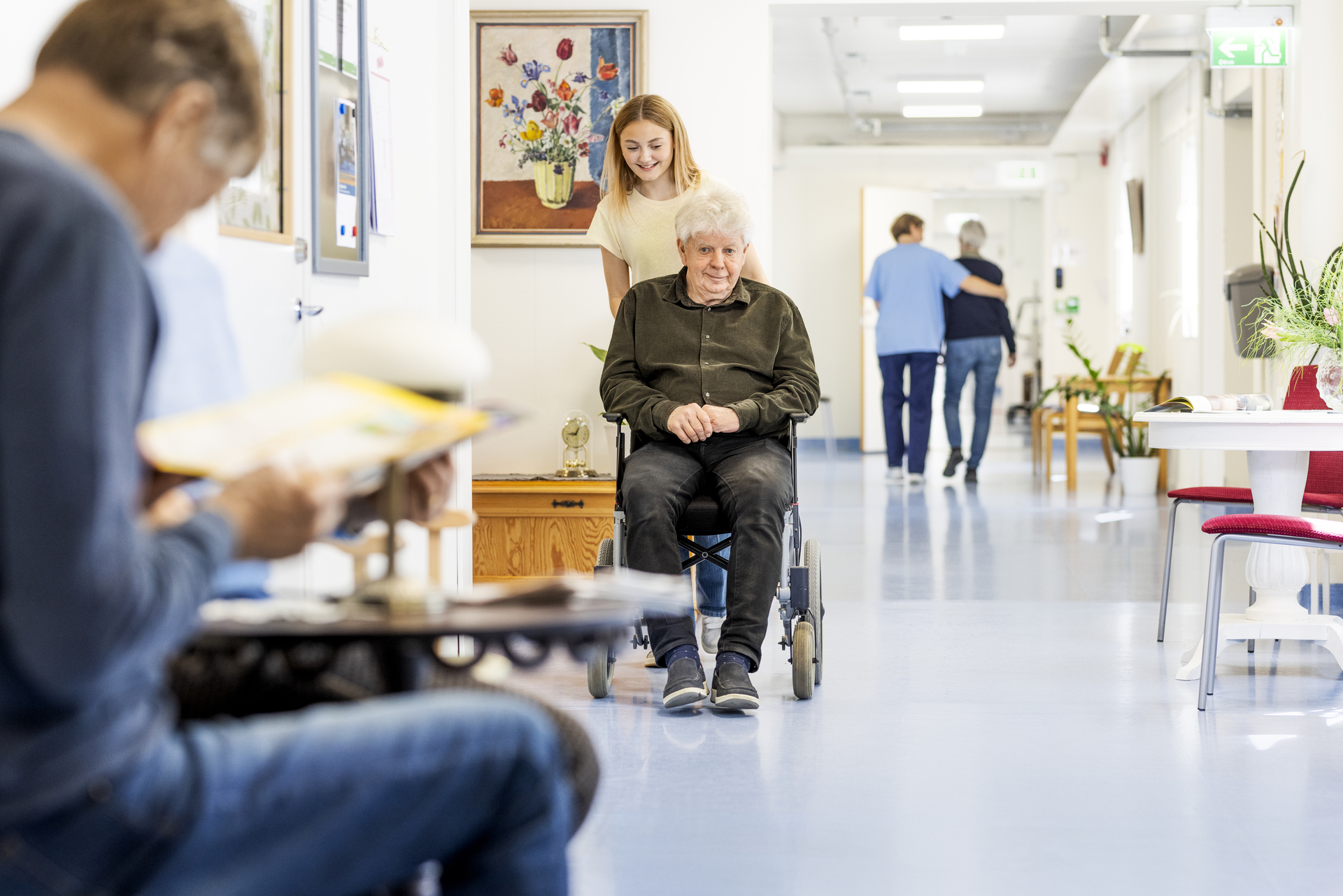 En boende i rullstol leds genom en korridor vid Borggården demensvård i Olofström. I miljön syns gemensamma utrymmen och personal som stöttar boende i vardagen, inom en modullösning från Expandia.