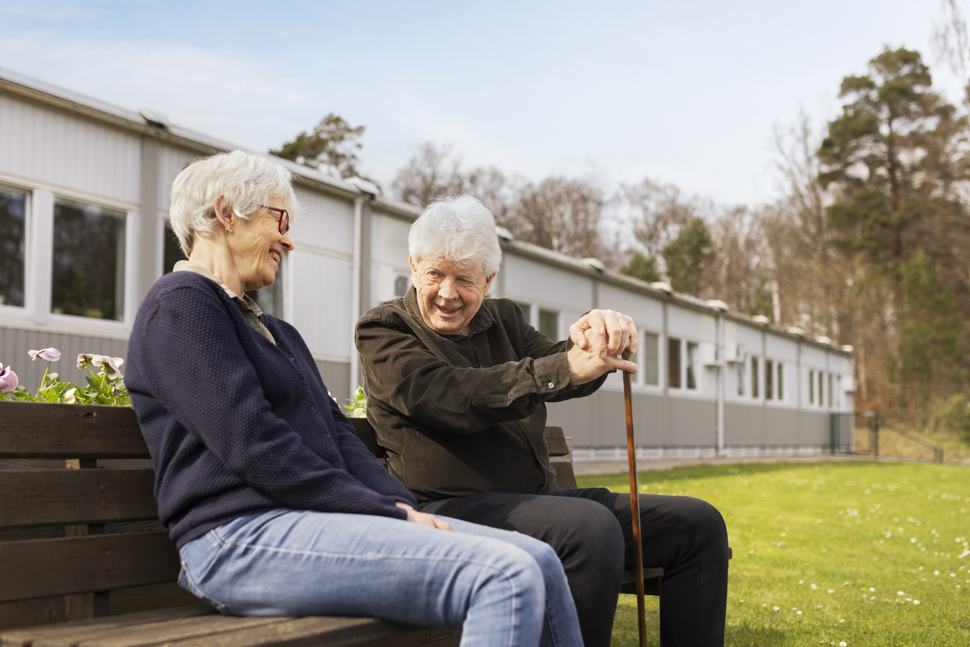 To beboere sitter på en benk i uteområdet ved Borggården demensomsorg i Olofström. I bakgrunnen sees en modulløsning fra Expandia som omgir gressområdet.