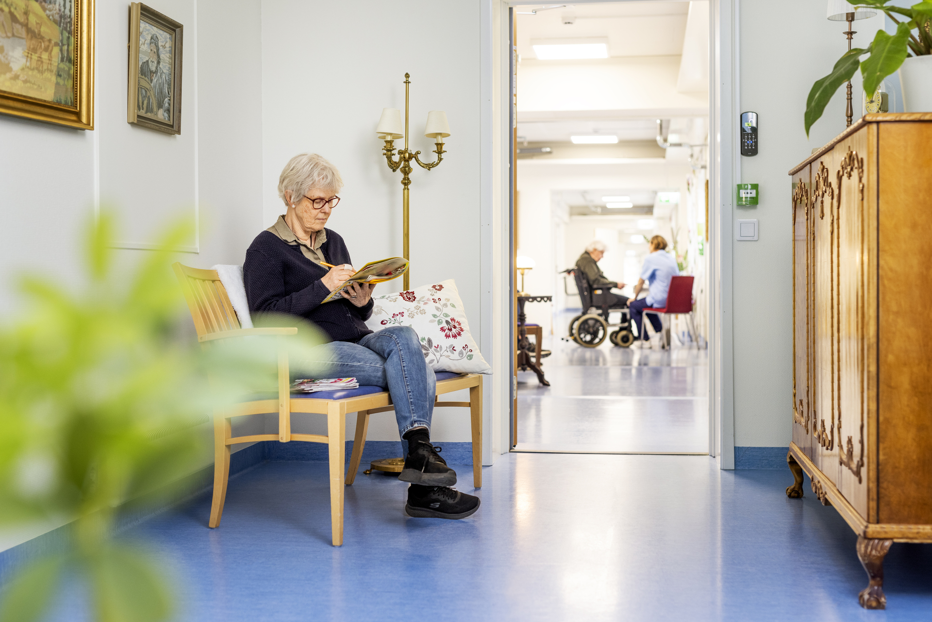En beboer sitter i en korridor og leser en avis ved Borggården demensomsorg i Olofström. I bakgrunnen sees en annen beboer og omsorgspersonell i samtale, i et lyst miljø som inngår i en modulløsning fra Expandia.