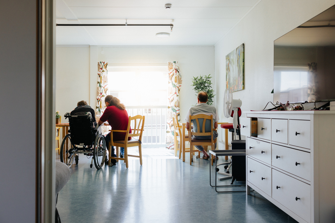 Gemensamt inomhusutrymme vid Herrhagsgården vård- och omsorgsboende i Falun där flera äldre personer sitter vid bord, varav en använder rullstol. Rummet har stora fönster som släpper in dagsljus samt möbler anpassade för gemenskap och vila.