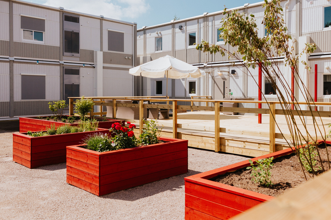 Innergård vid Herrhagsgården vård- och omsorgsboende i Falun med trätrall, ramp, parasoll och röda planteringslådor med blommor. Moduler i två plan ramar in gården och skapar en trivsam utemiljö.