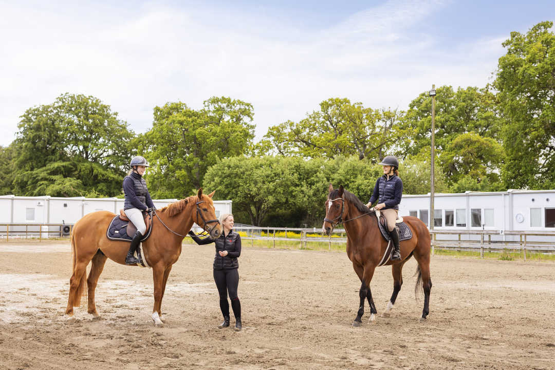 Två elever sitter till häst på en ridbana vid Sveriges Ridgymnasium i Svedala, tillsammans med en lärare som står mellan hästarna. I bakgrunden syns boendemoduler som används som elevboenden och är placerade intill skolområdet.