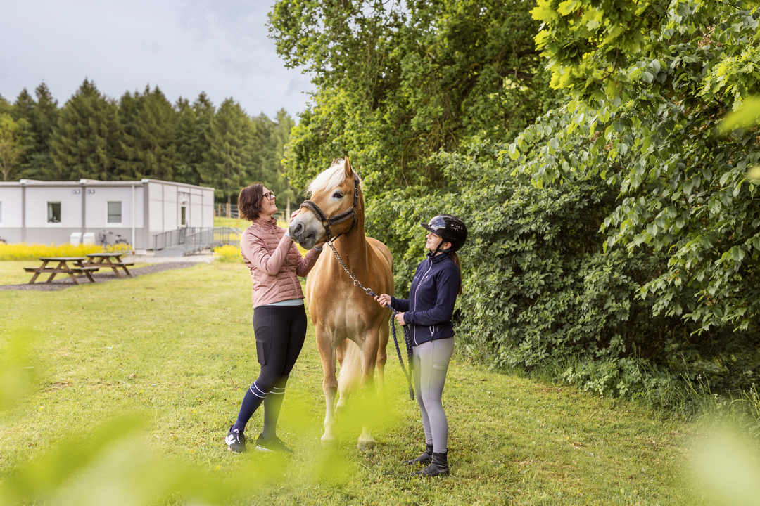 En elev och en lärare vid Sveriges Ridgymnasium i Svedala sköter om en häst på en gräsyta intill skolområdet. I bakgrunden syns boendemoduler som används som elevboenden, placerade i anslutning till verksamheten.