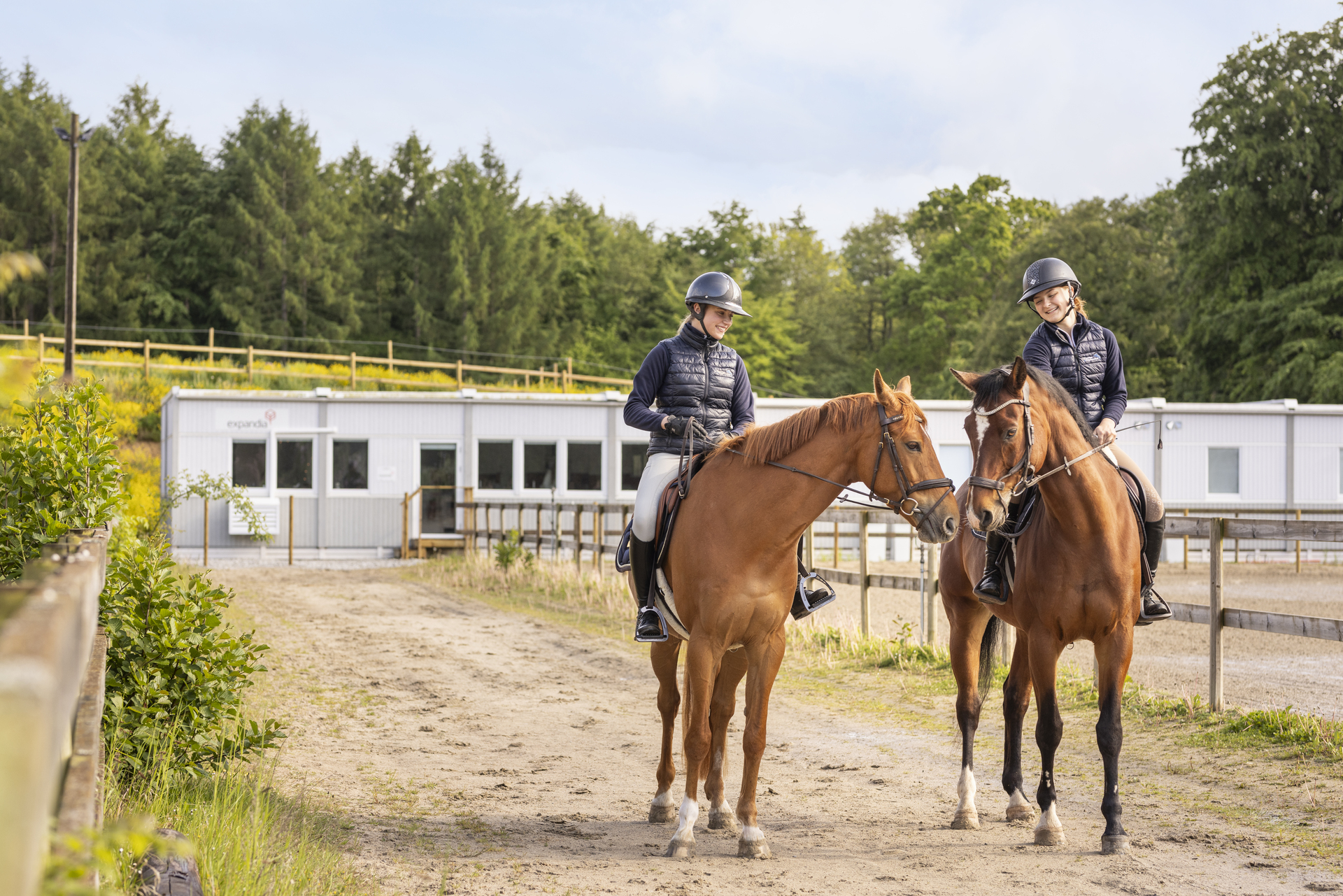 Två elever rider på hästar längs en ridväg vid Sveriges Ridgymnasium i Svedala. I bakgrunden syns boendemoduler som används som elevboenden och hyrs från Expandia, placerade intill skolområdet.