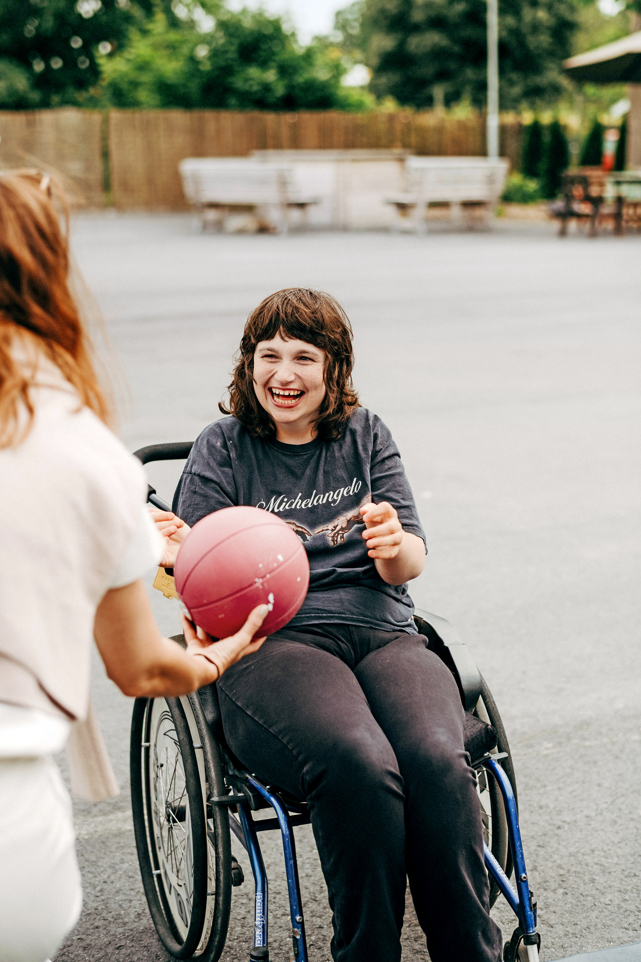 En person i rullestol ler og strekker seg etter en rød ball som blir gitt av en annen person under en felles aktivitet utendørs.